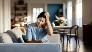 Une femme assise sur un canapé, tenant sa tête avec une expression de douleur sur son visage.